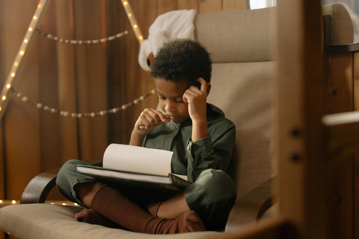 A boy curled up in a cozy chair, reading thoughtfully with his hand resting on his head