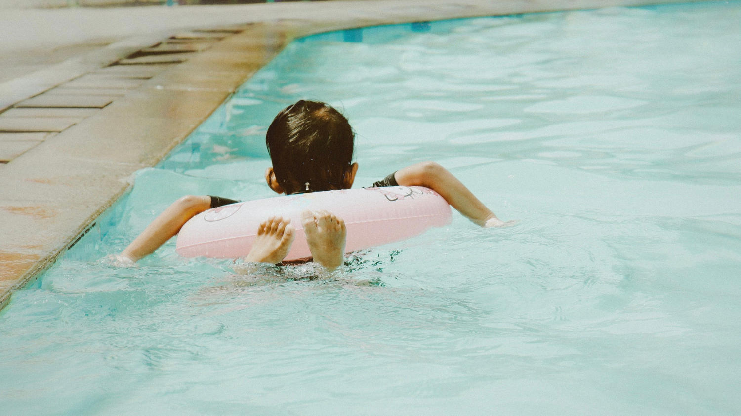 A kid floating on a pink pool ring in a swimming pool, seen from behind